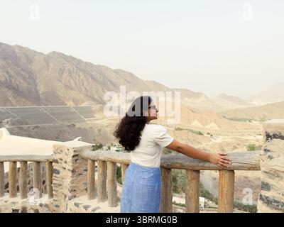 Eine Frau mit Blick auf die Berge, mit Sonnenbrille, weißem Hemd und blauem Rock. Sie steht an einem rustikalen Stein- und Holzgeländer Stockfoto