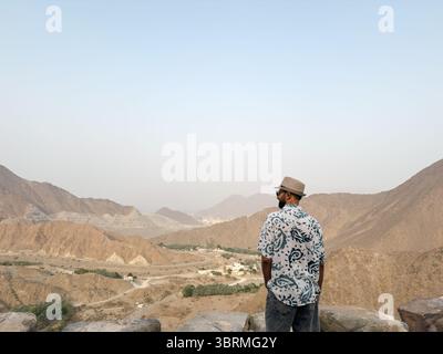 Ein Mann mit gemustertem Hemd und Hut, auf Felsen stehend, überblickt bergiges Gelände in einer unruhigen Landschaft Stockfoto
