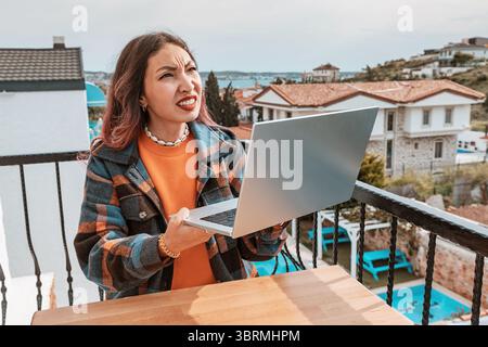 Eine junge frustrierte Frau, die aufblickte und schreite, während sie einen Laptop in der Hand hielt und vom Hotelbalkon aus aus aus der Ferne arbeitete Stockfoto