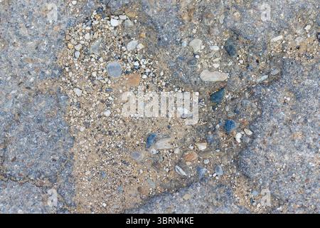 Nahaufnahme von beschädigtem Asphalt mit freiliegendem Kies und Sand. Die Textur ist rau und ungleichmäßig, mit Abweichungen in Farbe und Material. Stockfoto