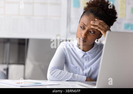 Der in Schwierigkeiten geratene afrikanische Büroangestellte sitzt am Schreibtisch mit Laptop Stockfoto