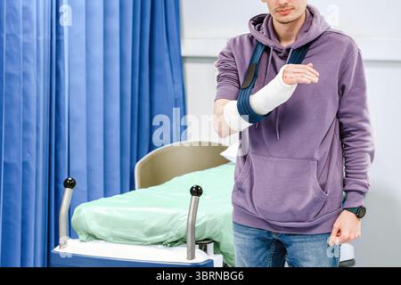 Ein junger Mann steht mit einem gebrochenen Arm in einem Guss, Arm ruht in einer Schlinge, Krankenhauszimmer Hintergrund mit einem leeren Krankenhausbett auf Rädern, blaue Privatsphäre-Knicke Stockfoto