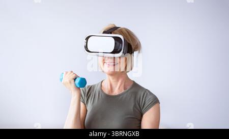 Sportliche reife Frau in VR-Headset arbeiten ihre Bizeps Mit Kurzhanteln während des inländischen Trainings Stockfoto