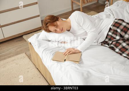 Hübscher junger müder Mann im Pyjama mit Kissen und Buch, der zu Hause auf dem Bett schläft Stockfoto