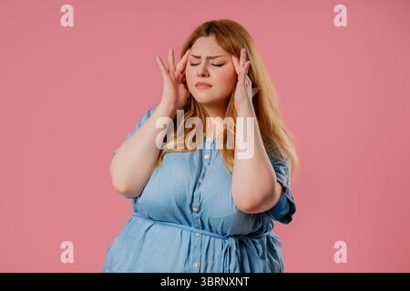 Frau in Übergröße, die Schläfe berührt, Kopfschmerzen hat, rosa Studio. Schmerzen. Stockfoto