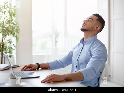 Erschöpfter junger Araber, der in einem modernen Büro neben dem Laptop sitzt Stockfoto