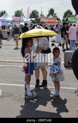 Bloomington, MN, USA. Juli 2025. Eine Frau mit zwei asiatischen Mädchen, die sich vor der Sommerhitze unter einem Schirm beim ausverkauften Panda fest Twin Cities schützen, das asiatische Kultur feiert. Das Panda fest ist eines der größten asiatischen Open-Air-Food-Festivals in den USA und bringt Hunderte authentischer asiatischer Speisen, Street Food und Kunstverkäufer auf den nördlichen Parkplatz der Mall of America. Quelle: Mariko Okada/Alamy Live News Stockfoto