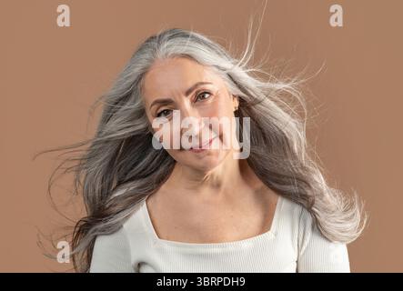 Porträt einer lächelnden älteren Frau, die mit langen und gesunden grauen, fliegenden Haaren auf braunem Studiohintergrund posiert Stockfoto