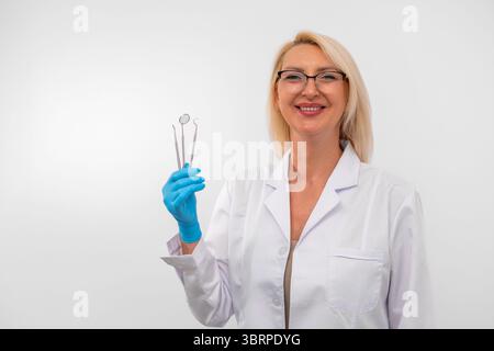 Blonde Zahnärztin mit Handschuhen und Laborkittel mit Zahninstrumenten. Reifer Zahnarzt in weißer Uniform mit Werkzeug, isoliert auf weiß. Das Konzept der Mundgesundheit, Hygiene und modernen Zahnheilkunde. Stockfoto