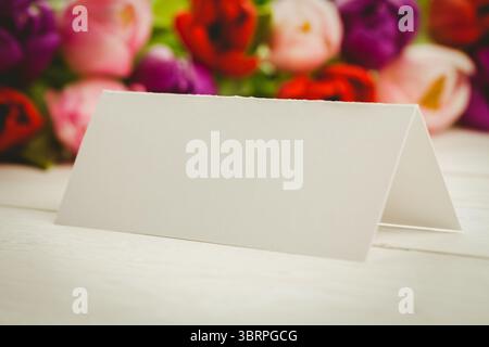 Flache leere Zelt-gefaltete Karte mit flachem Design sitzt auf einer Holzplatte mit verschwommenen Tulpenblüten dahinter Stockfoto