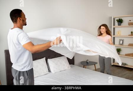 Hausarbeiten. Millennial multinationales Paar in der Liebe macht Bett zu Hause, Spaß haben, während Hausarbeit zusammen Stockfoto