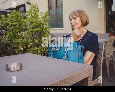Nachdenkliche Seniorin mit kurzen blonden Haaren und einer blauen Decke, die an einem Kaffeetisch im Freien sitzt, Hand am Kinn und mit einem grünen Busch im Hintergras wegblickt Stockfoto