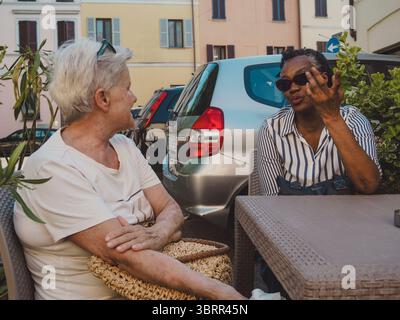 Zwei ältere Freundinnen, eine mit kurzen weißen Haaren und die andere mit Sonnenbrille und gestreiftem Hemd, genießen ein lebhaftes Gespräch in einem Café im Freien Stockfoto
