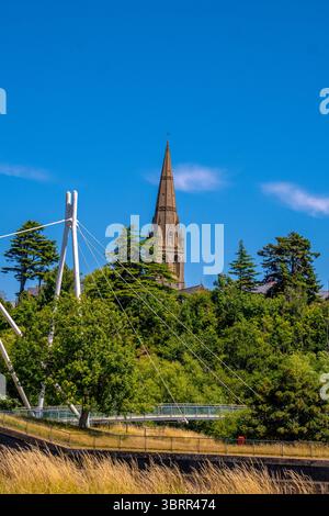 Millers überquert die Fußgängerbrücke über den Fluss exe in Exeter, Devon, mit einem Kirchturm, der an einem hellen Sommertag über den Baumkronen ragt Stockfoto