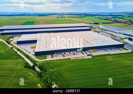 Großes modernes Lagerhaus mit Laderampen und geparkten LKWs, umgeben von grünen Feldern. Industrielogistik-Hub. Verkehrsinfrastruktur für den Bezirk Stockfoto