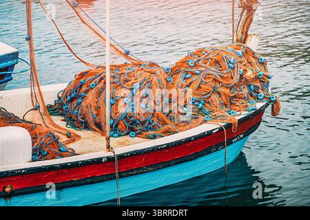 Fischernetze mit blauen Schwimmern liegen auf dem Bug eines kleinen, bunten Fischerbootes, die sanft auf dem ruhigen Wasser eines Hafens schwimmen Stockfoto