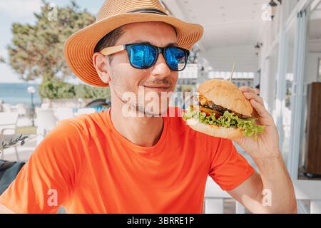 Junger Mann mit Sonnenbrille und Strohhut, der einen Burger in einem Restaurant im Freien mit Meerblick hält Stockfoto