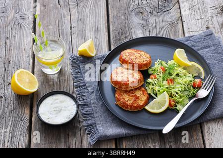 Geräucherte Lachsburger, Pasteten oder Kuchen serviert mit Krautsalat und Zitronenscheiben auf einem Teller auf rustikalem Holztisch mit Tzatziki-Sauce und Sprudelwasser Stockfoto