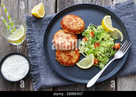 Geräucherte Lachsburger, Pasteten oder Kuchen serviert mit Krautsalat und Zitronenscheiben auf einem Teller mit Gabel auf rustikalem Holztisch mit Tzatziki-Sauce und Wasser Stockfoto