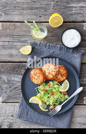 Geräucherte Lachsburger, Pasteten oder Kuchen serviert mit Krautsalat und Zitronenscheiben auf einem Teller auf rustikalem Holztisch mit Tzatziki-Sauce und Wasser, vertikal Stockfoto