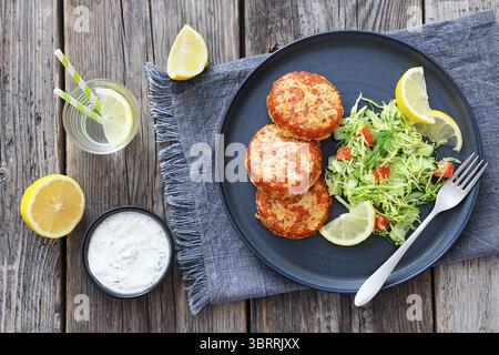 Geräucherte Lachsburger, Pasteten oder Kuchen serviert mit Krautsalat und Zitronenscheiben auf einem Teller auf rustikalem Holztisch mit Tzatziki-Sauce und Wasser, flach gelegt Stockfoto
