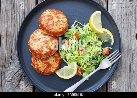 Geräucherte Lachsburger, Pasteten oder Kuchen serviert mit Krautsalat und Zitronenscheiben auf einem Teller mit Gabel auf rustikalem Holztisch, Draufsicht, flacher Lay, Nahaufnahme Stockfoto