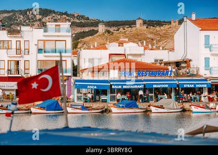 7. Mai 2025, Foca, Turkiye: Lebhafte Hafenszene mit farbenfrohen Booten, einer türkischen Flagge und einem Hügel mit historischen Windmühlen im Hintergrund Stockfoto