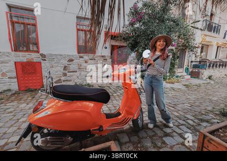 7. Mai 2025, Foca, Turkiye: Lächelnde Frau mit Hut neben einem orangen Vespa-Roller auf einer Kopfsteinpflasterstraße Stockfoto