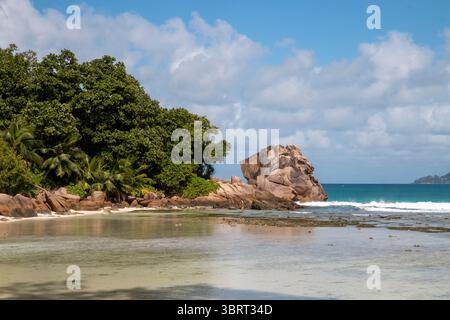 Eine ruhige Bucht mit einem großen Granitfelsen und üppigen Bäumen trifft unter einem bewölkten Himmel bei Anse Strong auf La Digue auf den Seychellen auf das sandige Ufer. Stockfoto