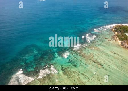 Ein Blick aus der Vogelperspektive zeigt die atemberaubende Küste der Seychellen, wo üppiges grünes Land auf das pulsierende türkisfarbene Meer und seine Korallenriffe trifft. Stockfoto