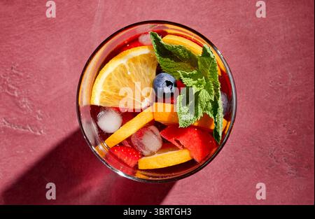 Nahaufnahme des roten Sangria-Glases mit frischen Obststücken, Eis und Beeren, dekoriert mit Minzblatt, Blick von oben, auf sonnigem hellem Hintergrund Stockfoto