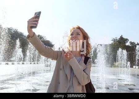 Junge kaukasische Frau, die ein lustiges Selfie-Foto mit ihrem Smartphone an den Wasserfontänen in einem sonnigen Stadtpark macht. Fröhliche Reiseerinnerungen festhalten, sh Stockfoto