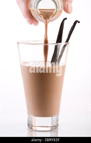Schokoladenmilch, die aus der Flasche in Glas mit Vanille auf weißem Hintergrund gegossen wird Stockfoto