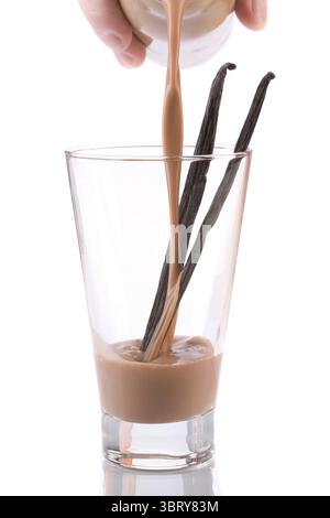 Schokoladenmilch, die aus der Flasche in Glas mit Vanille auf weißem Hintergrund gegossen wird Stockfoto