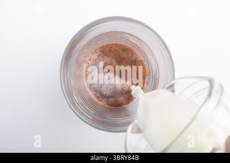 Zubereitung von Kakaogetränk im Glas, Hinzufügen von Milch zu Kokaenergie von oben auf weißem Hintergrund Stockfoto