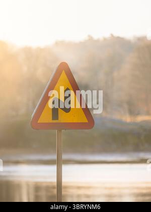 In Schweden steht ein leuchtend farbiges Warnschild hoch, das die Fahrer auf eine bevorstehende scharfe Kurve aufmerksam macht. Das sanfte Morgenlicht verstärkt die ruhige Atmosphäre Stockfoto