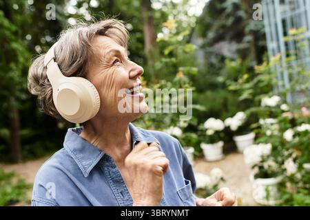 Eine Seniorin mit Kopfhörern tanzt fröhlich zwischen blühenden Blumen in einem üppigen Garten. Stockfoto