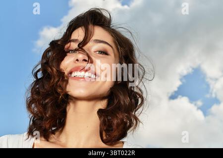 Eine fröhliche junge Frau mit gewellten braunen Haaren lächelt hell und genießt den Sommertag im Freien. Stockfoto