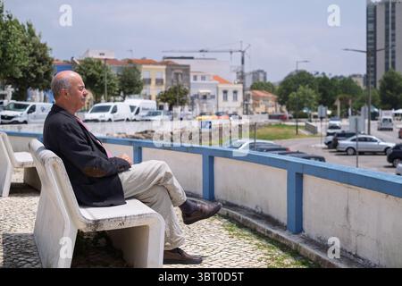 Ein älterer Spanier in einem Blazer sitzt auf einer Bank in einer kleinen portugiesischen Stadt und blickt auf den Strand Stockfoto