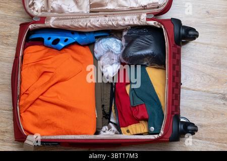 Offener roter Koffer mit bunten Kleidern, blauen Sandalen und verpackten Gegenständen auf einem Holzboden. Konzept der Reisevorbereitung Stockfoto