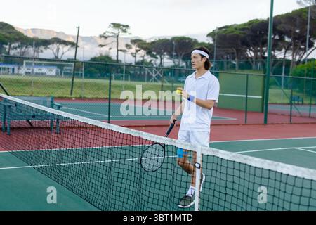 Asiatischer Mann, der in der Nähe des Netzes auf dem Hartplatz steht und Schläger mit Ball in weiß hält, Kopierraum Stockfoto