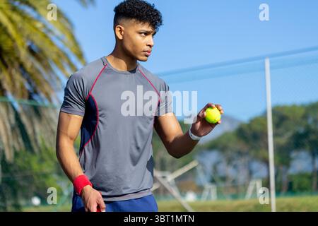 Afroamerikaner, der in grauem Hemd auf dem Tennisplatz steht und einen Tennisball hält, der den Serviertisch vorbereitet Stockfoto