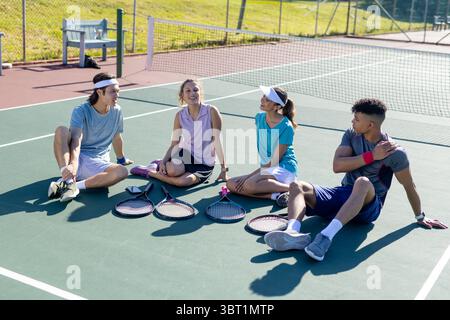 Verschiedene Freunde sitzen auf dem Tennisplatz und unterhalten sich mit Schlägern und einer Bank neben dem Maschendrahtzaun Stockfoto