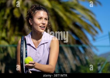 Frau mit violettem Polotop, die auf dem Tennisplatz steht und Schläger und Ball am Kettenzaun hält Stockfoto