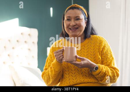 Reife Frau, die einen Senfpullover trägt und einen Keramikbecher hält, während sie im hellen Schlafzimmer auf dem Bett sitzt Stockfoto