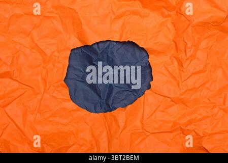 Eine kreisförmige Öffnung aus zerknittertem orangefarbenen Papier, die einen Hintergrund aus dunkelblauem, ähnlich gefaltetem Papier freigibt. Stockfoto