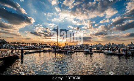 Malerischer Sonnenuntergang über dem Hafen mit verstreuten Wolken und Sonnenstrahlen, die durch die Wolken kommen, wunderschöner Blick auf den Hafen von Dubai mit Bootssteg und geparkten Booten Stockfoto