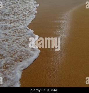 Nahaufnahme einer sanften Meereswelle mit weißem Schaum, der über goldenem Sand an einem Strand in Sardinien, Italien, wäscht. Ruhige Küstenszene unter natürlichem Sonnenlicht, pro Stockfoto