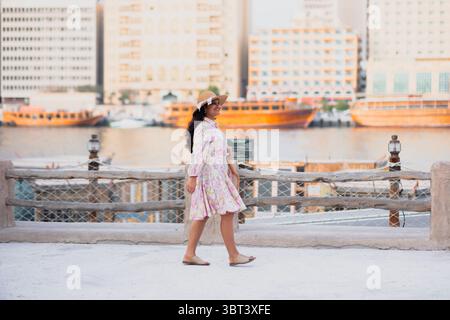 Junge Frau in ethnischer Kleidung und Hut, die Al Seef Touristenziel in Dubai erkundet. Mit traditioneller emiratischer Architektur und Abra-Booten genießt sie den Charme Stockfoto
