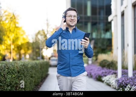 Ein Mann in einem blauen Hemd hört Musik auf Kopfhörern, während er draußen mit seinem Telefon läuft. Stockfoto